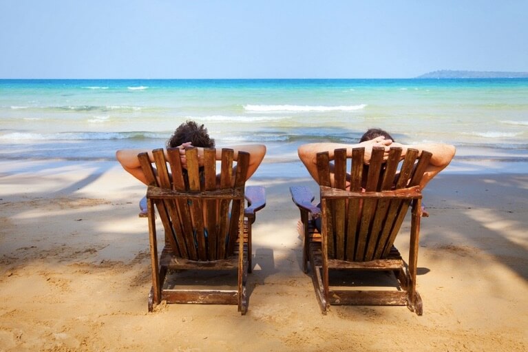 Zwei Männer sitzen auf Holzliegen am Strand und schauen aufs Meer