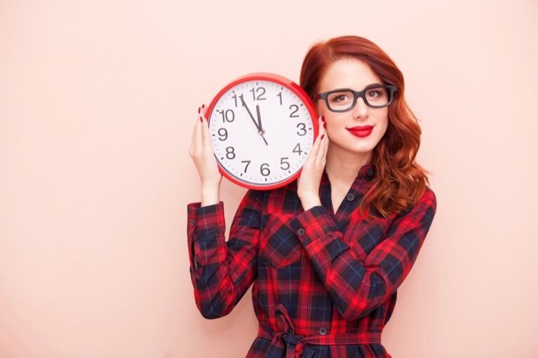 Rothaarige Frau mit Brille hält eine runde Wanduhr in der Hand